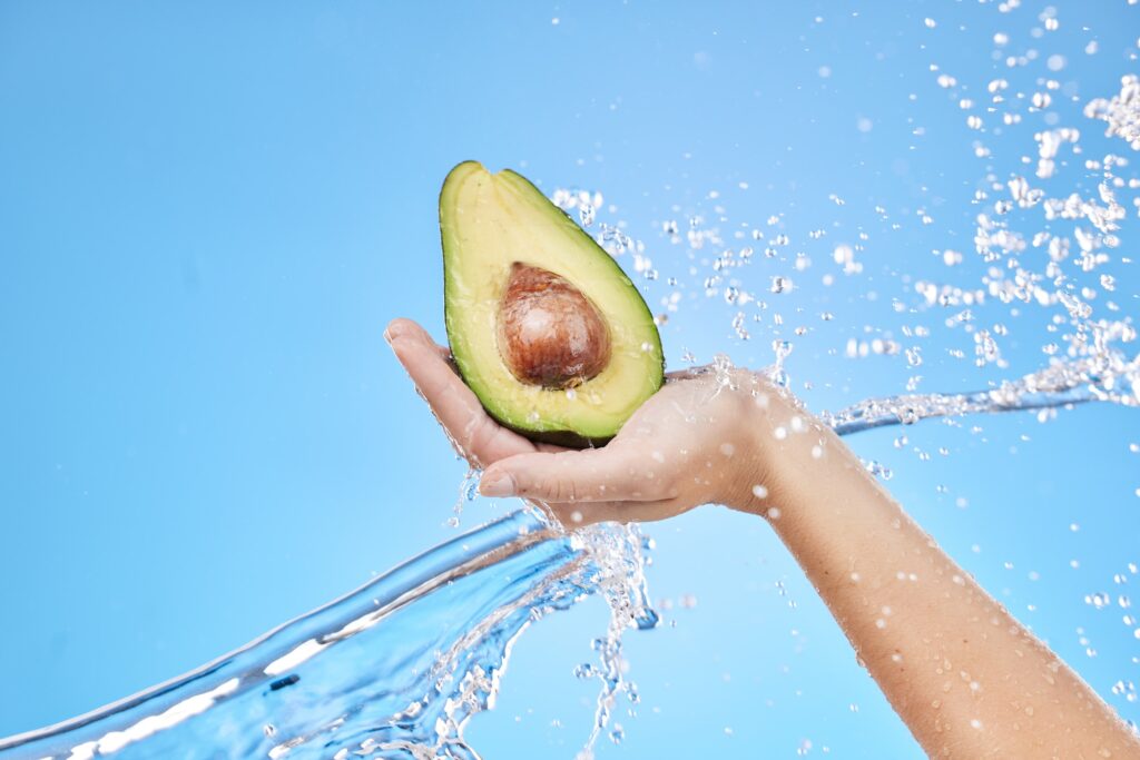 Hand, water and avocado for beauty splash and organic skincare product on a blue studio background.